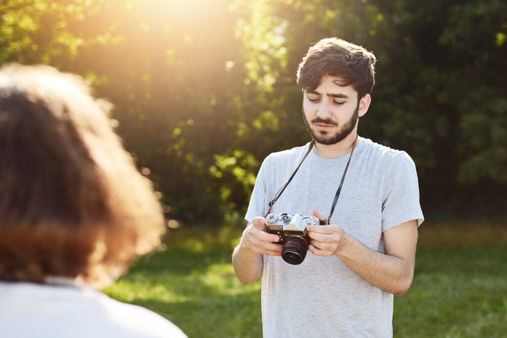 Attractive male with beard and stylish hairdo making photos of his girlfriend who is posing at nature looking pictures which he got at his retro camera. Professional photographer doing his work